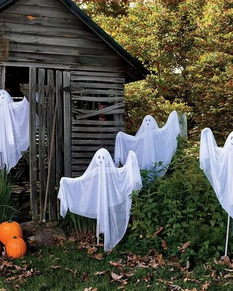 fantômes en voile blanc plantés dans un jardin devant une cabane en bois et des citrouilles orange