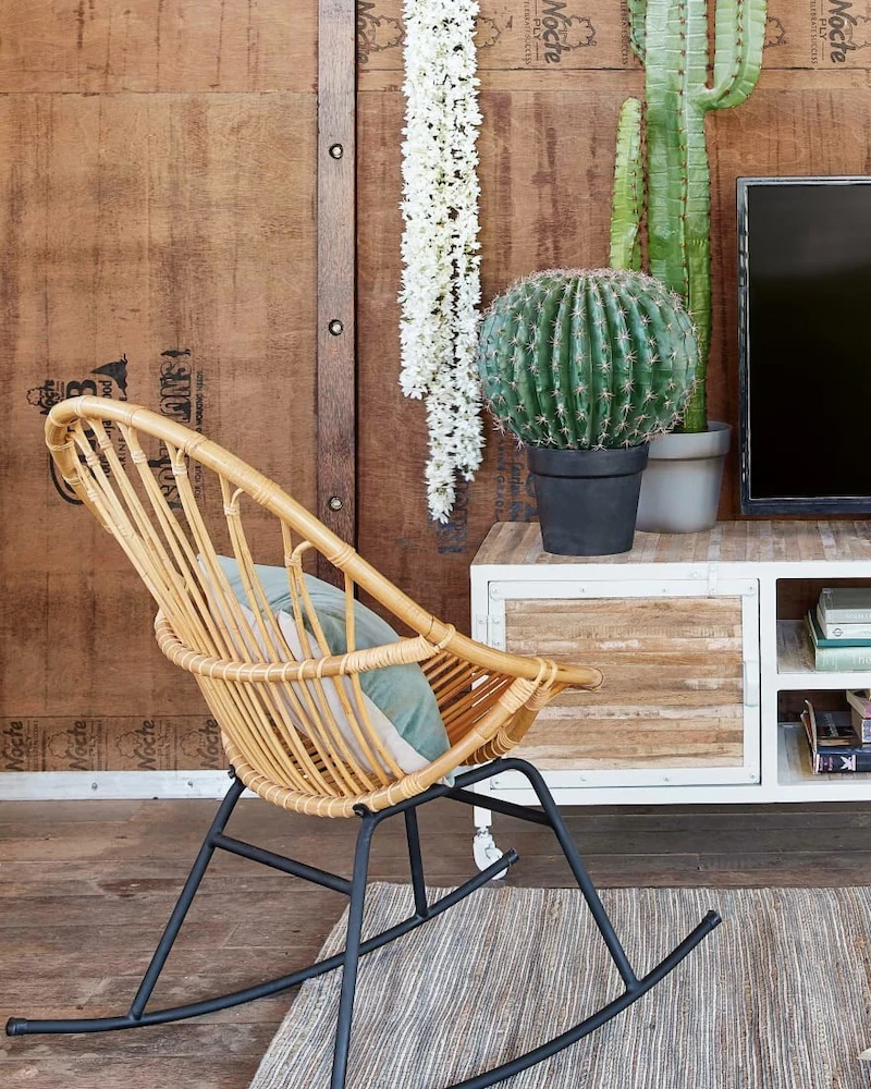 Fauteuil à bascule Petunia en rotin couleur miel sur piètement en acier noir devant un meuble TV