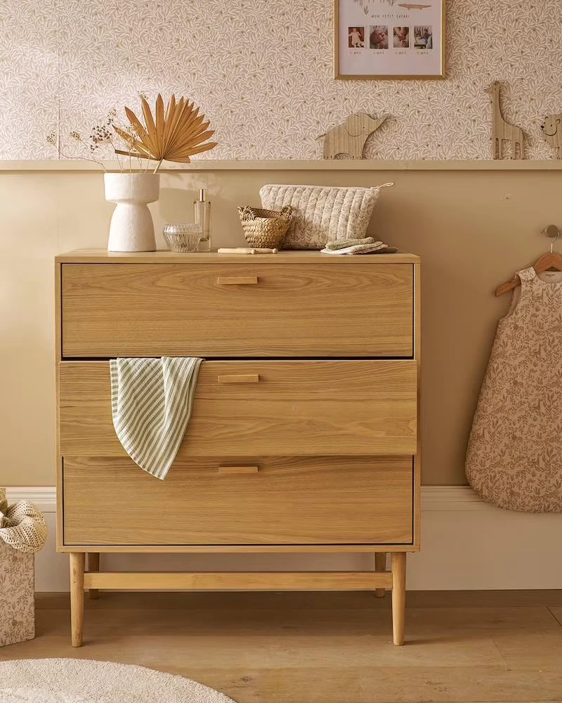 Commode bébé en bois clair à trois tiroirs sur piètement en bois devant un papier peint fleuri