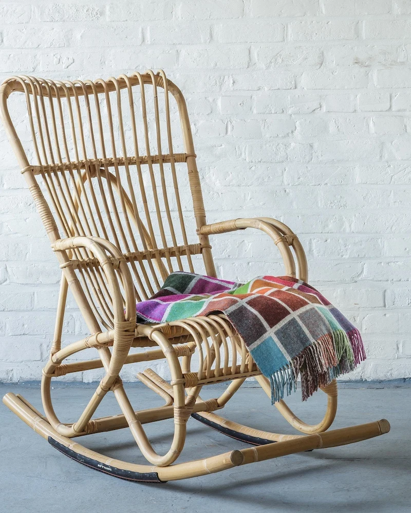 Rocking chair Marlène de KOK en rotin naturel à haut dossier avec plaid coloré devant un mur de briques blanches