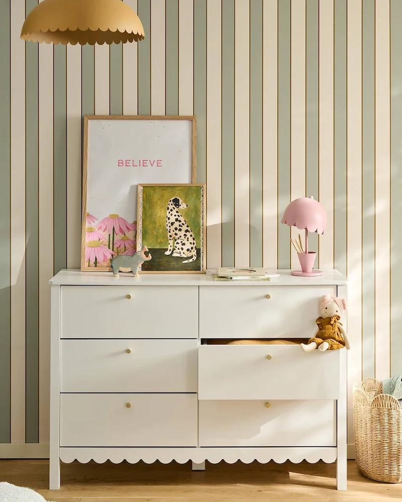 Grande commode bébé blanche à six tiroirs avec bas festonné devant un papier peint rayé