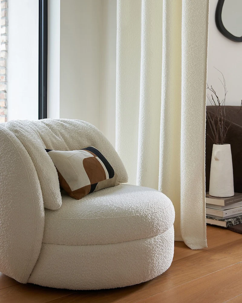 Fauteuil pivotant Rondaya en bouclette écrue au dossier enveloppant près d'un rideau blanc