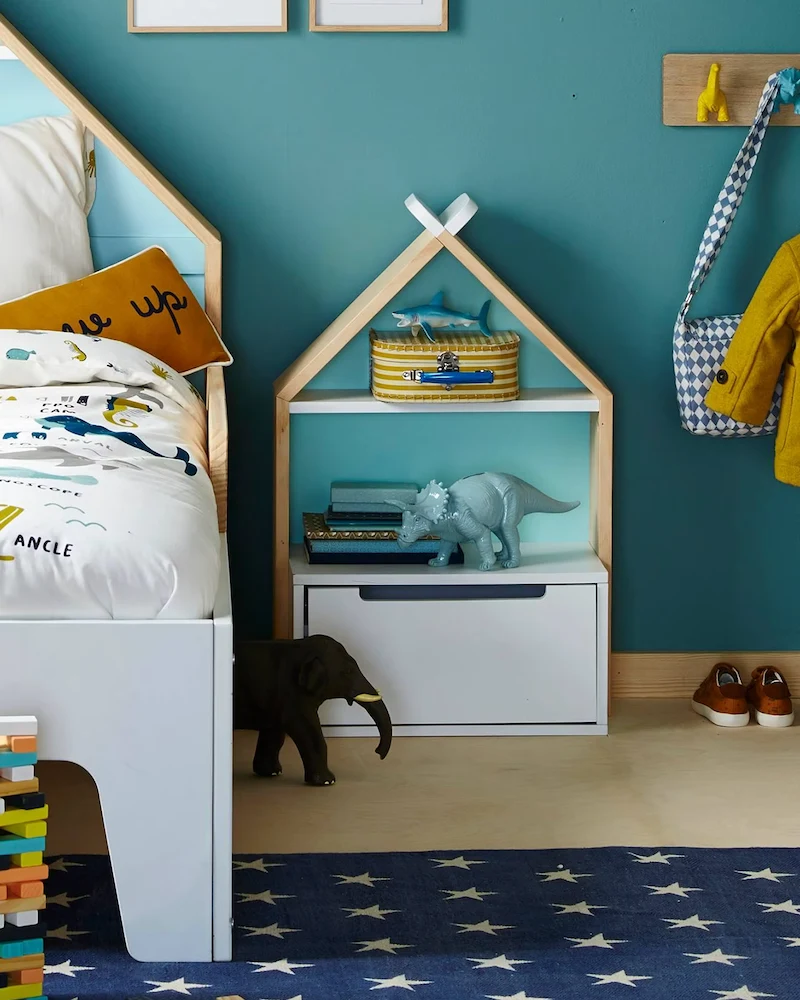 Table de chevet enfant en forme de maisonnette avec niche et compartiment inférieur devant un mur bleu canard