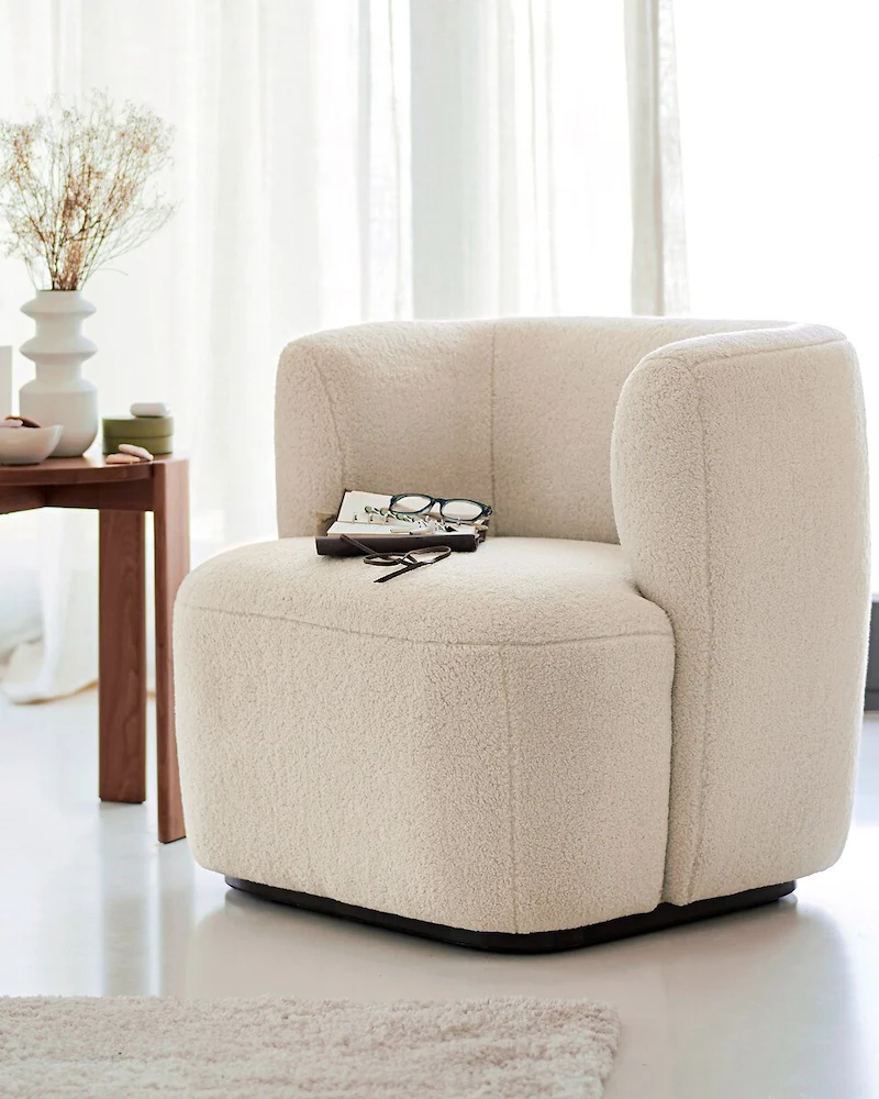Fauteuil Nidou en bouclette beige épaisse aux formes arrondies devant une fenêtre
