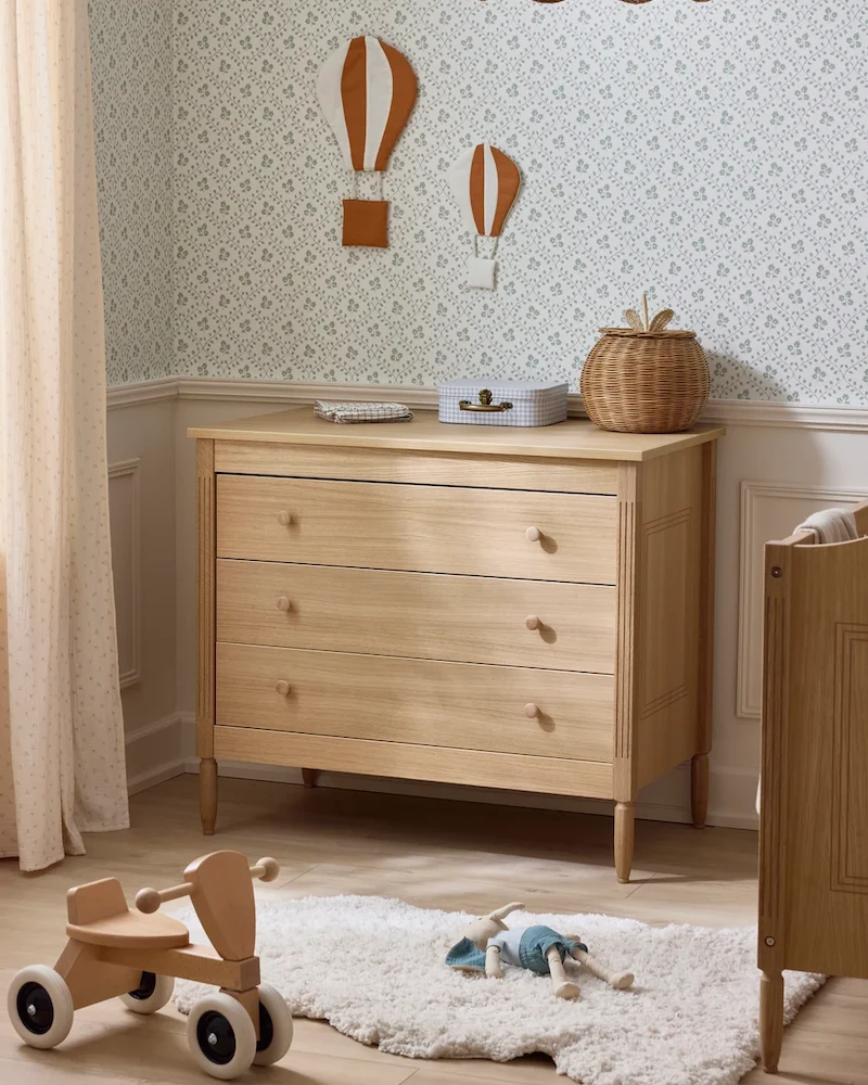 Commode bébé en bois clair à trois tiroirs sur pieds fins devant un papier peint à motifs
