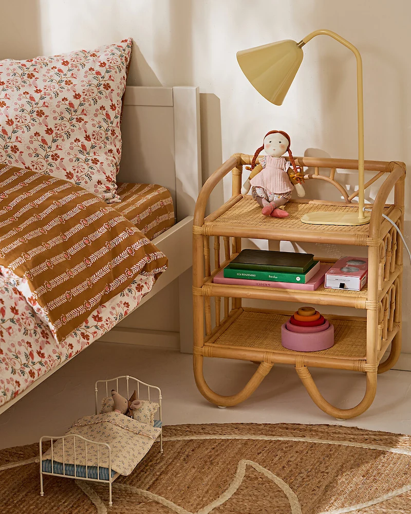 Table de chevet enfant à l'aspect rotin avec plusieurs niveaux ouverts et base arrondie près d'un lit au linge fleuri