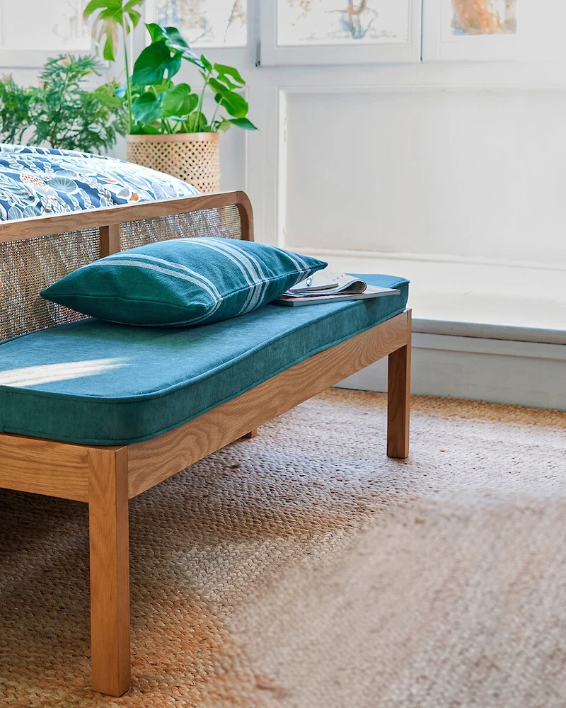 Banc Scillia en chêne avec dossier en cannage et coussin velours bleu canard au pied d'un lit