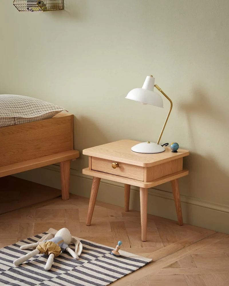 Table de chevet enfant à l'aspect bois clair avec un tiroir sur pieds fins et une lampe blanche