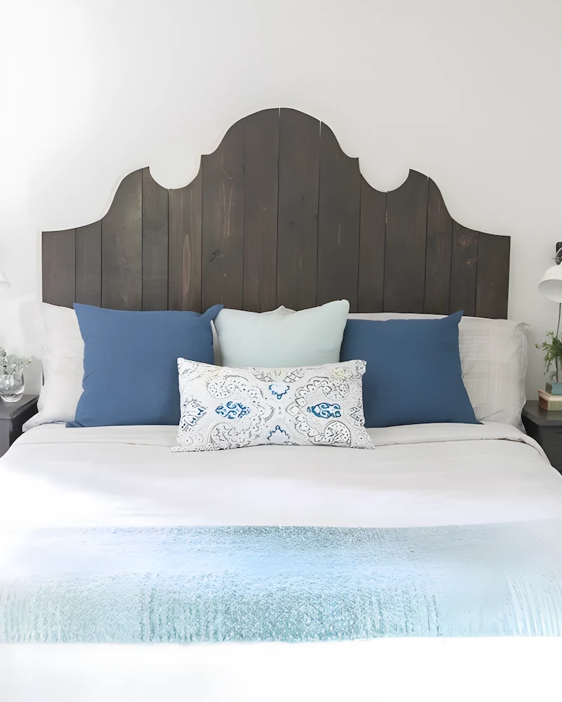 Tête de lit en planches de bois foncé à la silhouette chantournée derrière un lit blanc et coussins bleus