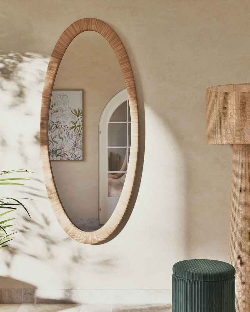 Grand miroir ovale Sariel en rotin tressé posé contre un mur clair près d'un pouf vert