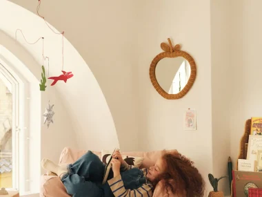 Enfant qui lit sur un pouf rose dans une chambre Ferm Living avec miroir pomme en rotin et bibliothèque Rivan