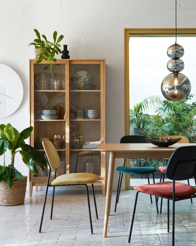 Salle à manger scandinave avec table en bois clair, chaises dépareillées, vaisselier et grandes plantes
